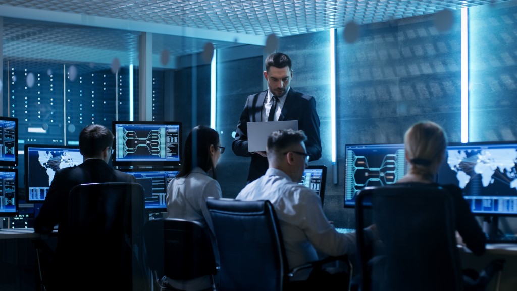 Professional IT Engineers Working in System Control Center Full of Monitors and Servers. Supervisor Holds Laptop and Holds a Briefing on Network Infrastructure Security