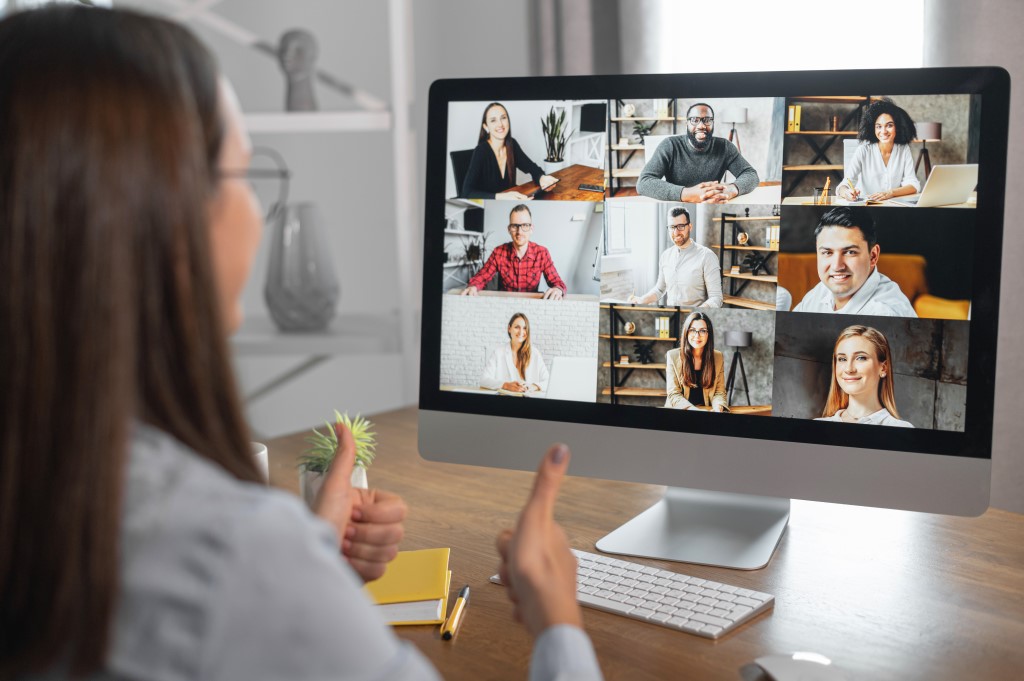 Businesswoman seats at workplace, talking online and showing thumbs up to a diverse team, a lot of multi-ethnic people on the monitor. Online video conference, morning meeting on the distance, video meeting with diverse group of people.